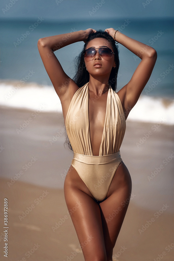 A slender and tanned brunette model in sunglasses and a gold bikini touches her hair. Luxury asian woman on the beach - obrazy, fototapety, plakaty 