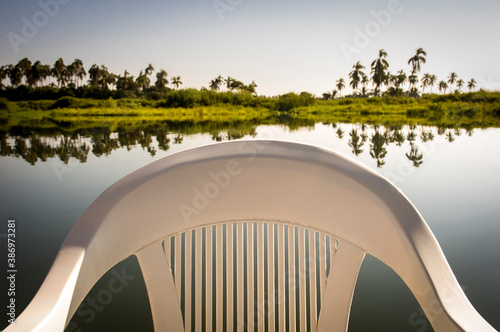 Close-up of an empty white plastic stackable chair with arms, and in the background the lush Coyuca de Benitez lagoon near Acapulco, in the Mexican state of Guerrero photo
