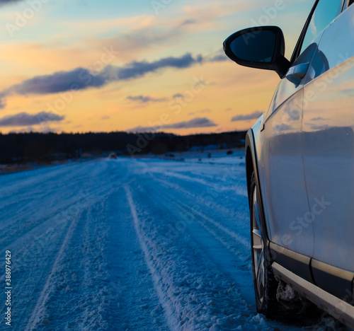 The left wheel and door of a white car on a snowy road during sunset with winter reflections on a frosted paint and copy space on the left