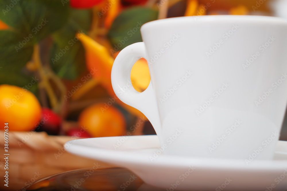 White porcelain conical cup with an eyelet on a saucer close-up. A cup on the background of a basket with a bouquet of imitation of branches of autumn trees with yellowed leaves. White porcelain conical cup with an eyelet on a saucer close-up. A cup on the background of a basket with a bouquet of imitation of branches of autumn trees with yellowed leaves.
