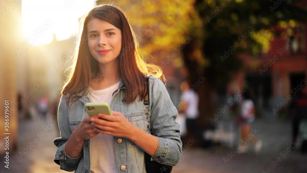 Happy young beautiful female student in stylish clothes texting message on smartphone on city street in slow motion Happy young beautiful female student in stylish clothes texting message on smartphone on city street in slow motion