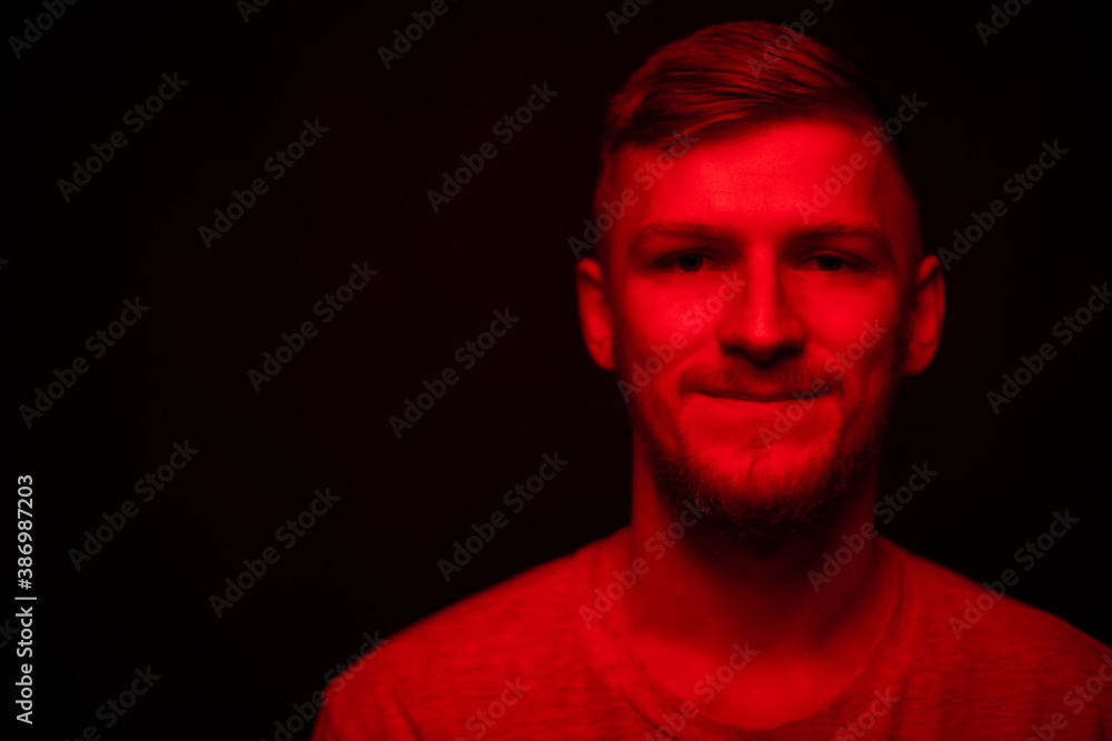 Low key portrait of bearded young confident man full-face looking at camera red light on black backdrop copy space. Layout for ads social assistance, call line for singles. Stress management concept Low key portrait of bearded young confident man full-face looking at camera red light on black backdrop copy space. Layout for ads social assistance, call line for singles. Stress management concept