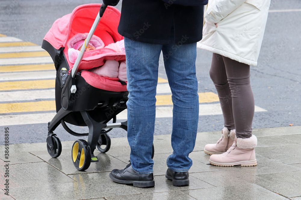 Fototapeta premium Couple with a kid in baby stroller standing in front of pedestrian crossing. Concept of road safety, parents with child in autumn city