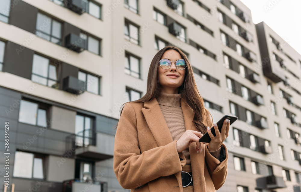 Obraz premium Business woman curly woman wearing trendy sunglasses walks down the central city street and uses her phone. Pretty summer woman in brown jacket walks down the street looking at her mobile phone.