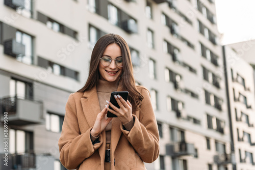 Happy pretty brunette girl using mobile phone near office, beautiful woman browsing phone and doing online shopping in a mobile app while smiling walking near shopping mall.