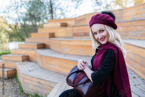 A smiling woman sits on a summer theater bench, made of wood in a burgundy coat and biret, an adult looks at the camera, in the fall against a background of blue clouds.