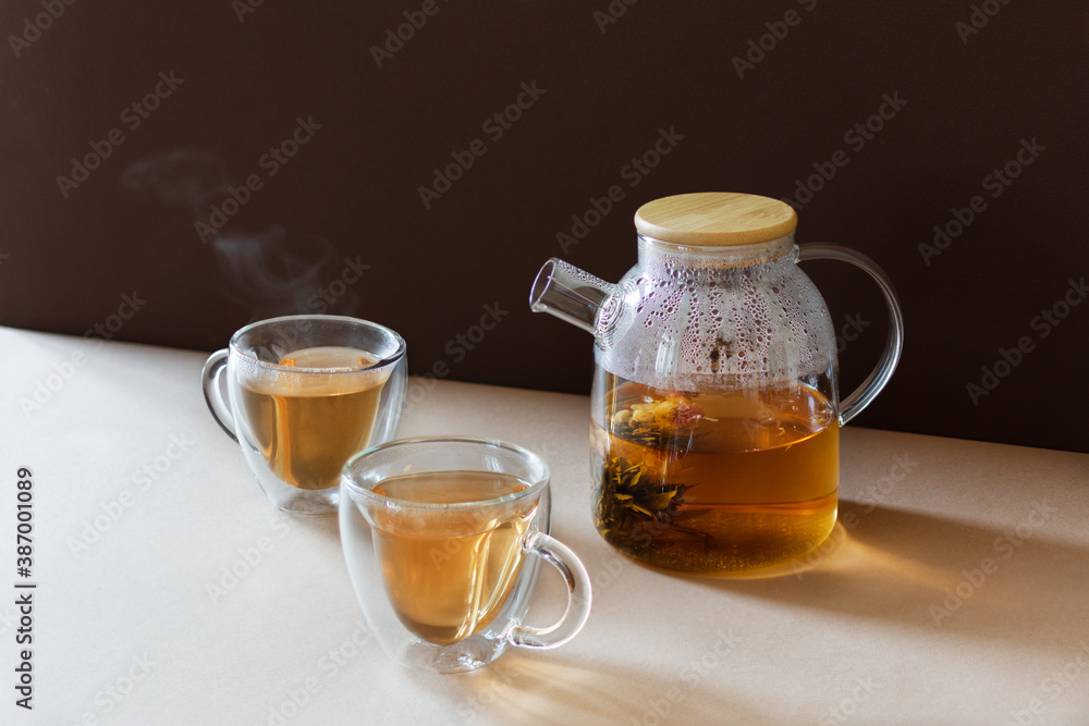 Glass tea pot with flower blooming tea and two tea cups on brown modern background.