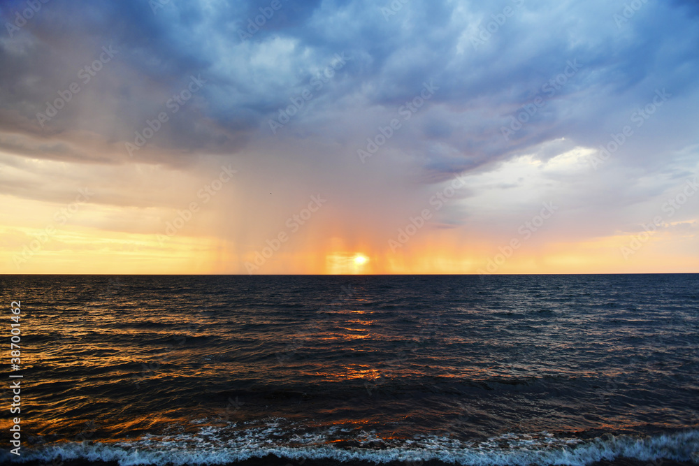 Thundercloud and streams of rain against the setting sun over the Mediterranean Sea. Foamy surf on the shore. Tropical downpour. Seascape.