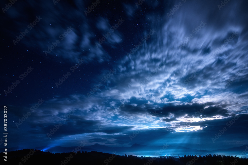 Beautiful night landscape with clouds and stars in the sky against the background of mountains with forests. Moon illuminated lanscape.