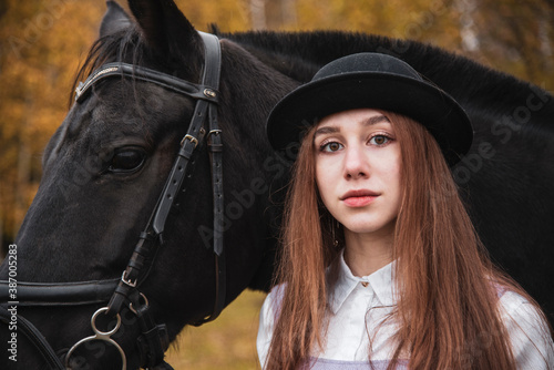 Red-haired girl in a black hat with a horse in the autumn forest.