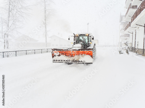 A snow plow tractor drives on a snowy road along a building during a severe blizzard