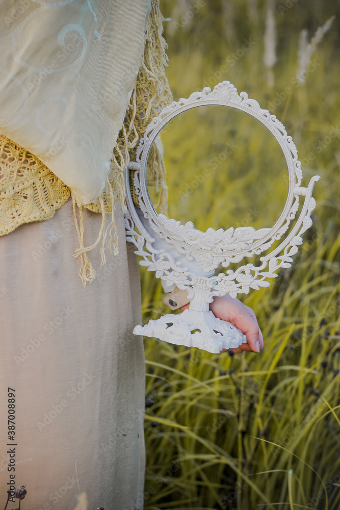 Fototapeta premium A Muslim woman stands on the field and holds a mirror in her hands. Muslim woman in beige hijab. Beauty, femininity.