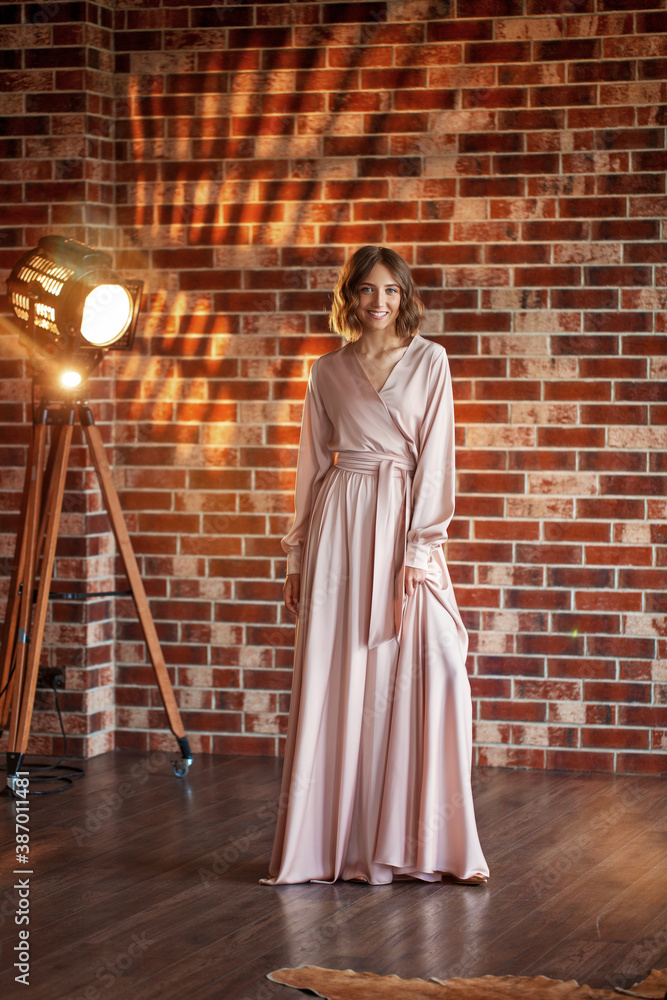  Beautiful young brunette in long beige dress posing outdoor near red brick wall