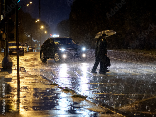 View of a pedestrian crossing in the city at night during a heavy downpour. Silhouettes of people with umbrellas in the headlights of the car. People cross the road. 