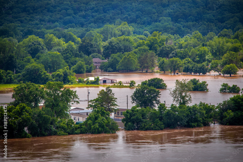 Muddy flood water swirls around a tree line and trailer houses and a barn with an upscale house in the distance under a forested hill where a dike has failed