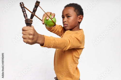 Canvas-taulu Isolated image of serious concentrated little black boy holding Y-shaped stick with elastic, shooting green apple, having focused facial expression
