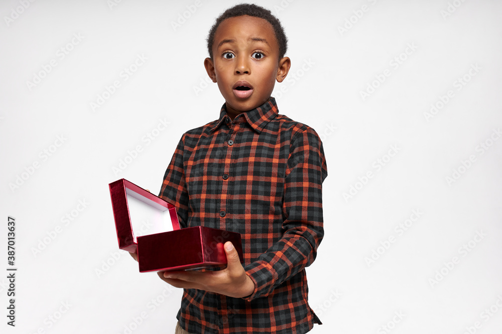 Studio shot of funny surprised little African boy in checkered shirt keeping mouth wide holding opened box with best birthday present, can not believe his eyes being excited with what he sees inside