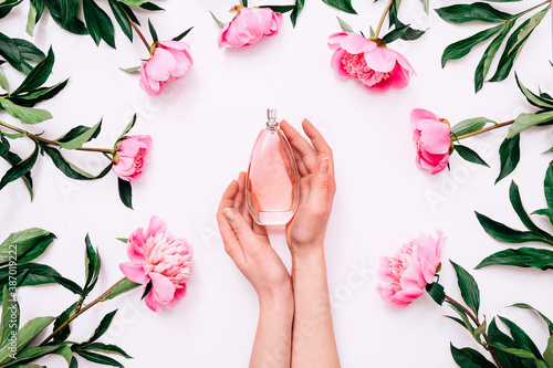 Pink bottle of women's perfume in female hands and peony flowers around on white background. Spring gentle fragrance for women. Top view, flat lay