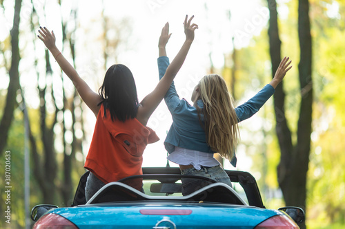Back view of a gorgeous pretty young two women friends driving the car with raised hands.