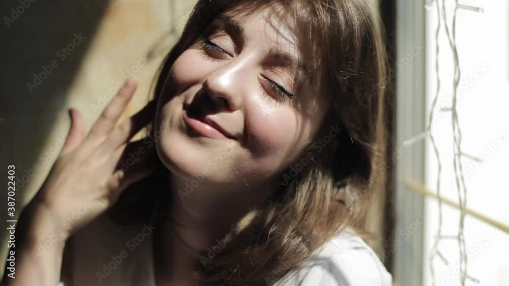 A tender young woman in a white T-shirt sits against the background of a window with garlands on a summer day and looks at the camera smiling. Front view. Close-up A tender young woman in a white T-shirt sits against the background of a window with garlands on a summer day and looks at the camera smiling. Front view. Close-up