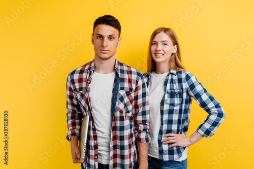 Beautiful young couple of students, man and woman with laptop on yellow background