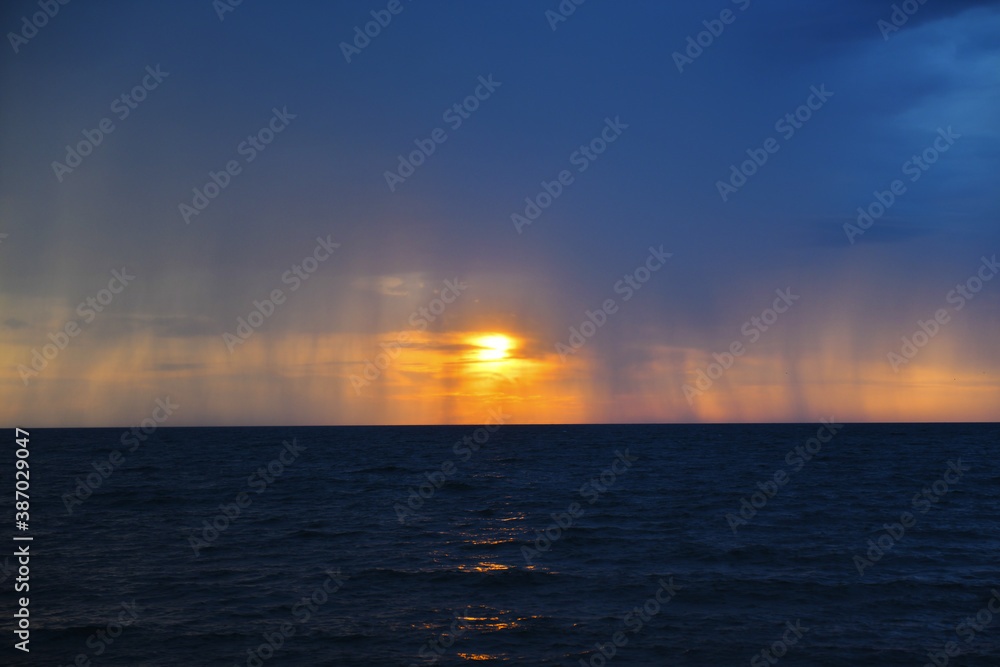 Thundercloud and streams of rain against the setting sun over the Mediterranean Sea. Foamy surf on the shore. Tropical downpour. Seascape.