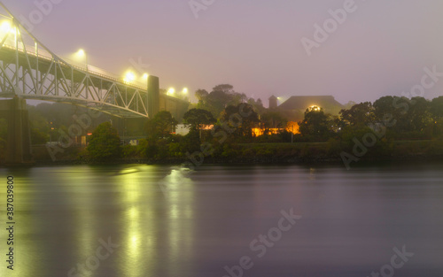 Foggy and Rainy Moody Night Landscape under Sagamore Bridge at Cape Cod Canal. Lighted Village and Yellow and Orange Light Reflections on the Water.