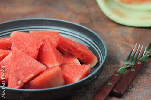 Buah Semangka or watermelon fruits, served on ceramic grey plate with fork beside, brown grainy background. Selective focus, close up. 