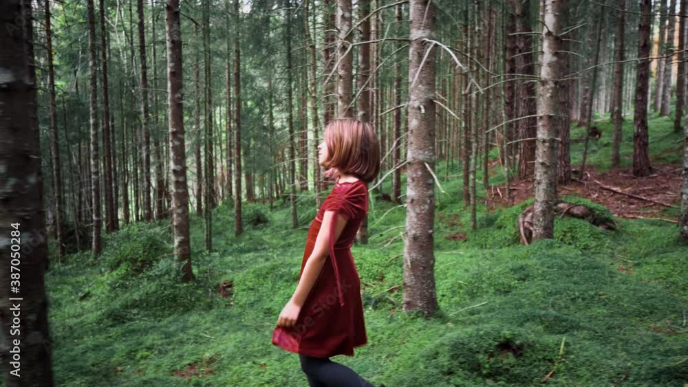 Truck shot of a girl with short hair in a red dress walking through a dense spruce forest, looking curiously around herself. Truck shot of a girl with short hair in a red dress walking through a dense spruce forest, looking curiously around herself.