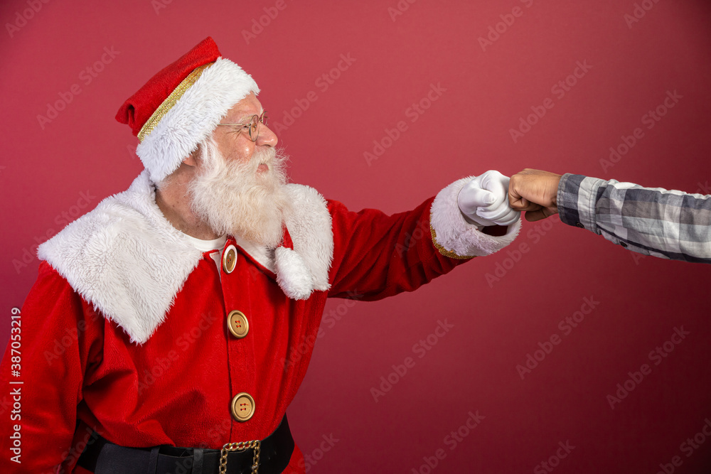 Real Santa Claus with red background, wearing glasses and hat doing elbow salute. Christmas with social distance. Covid-19 - obrazy, fototapety, plakaty 