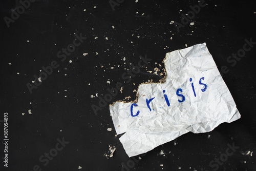 White crumpled burnt sheet of paper with word "Crisis" on black background with dispelled ash. Copy space. Top view. Global world economy collapse concept