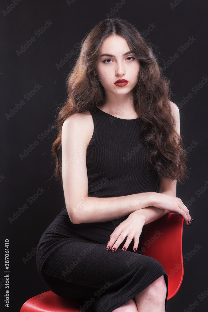 Beautiful slender young brunette with bright makeup in a black dress on a red chair Beautiful slender young brunette with bright makeup in a black dress on a red chair