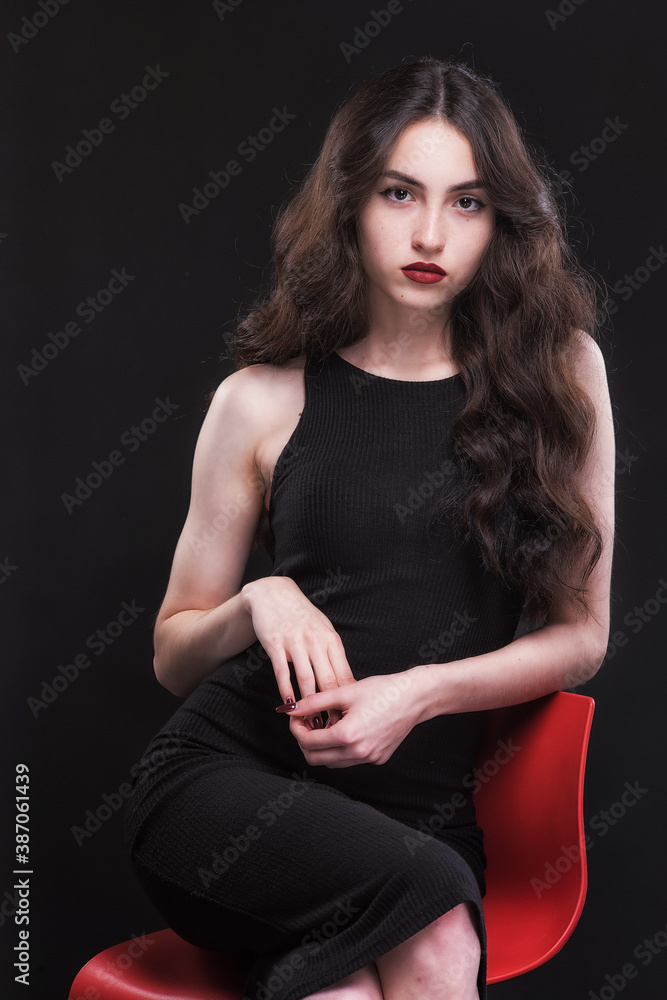 Beautiful slender young brunette with bright makeup in a black dress on a red chair Beautiful slender young brunette with bright makeup in a black dress on a red chair