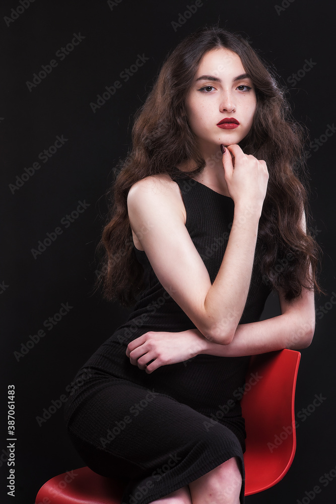 Beautiful slender young brunette with bright makeup in a black dress on a red chair Beautiful slender young brunette with bright makeup in a black dress on a red chair