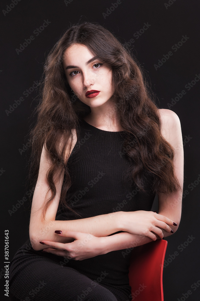 Beautiful slender young brunette with bright makeup in a black dress on a red chair Beautiful slender young brunette with bright makeup in a black dress on a red chair
