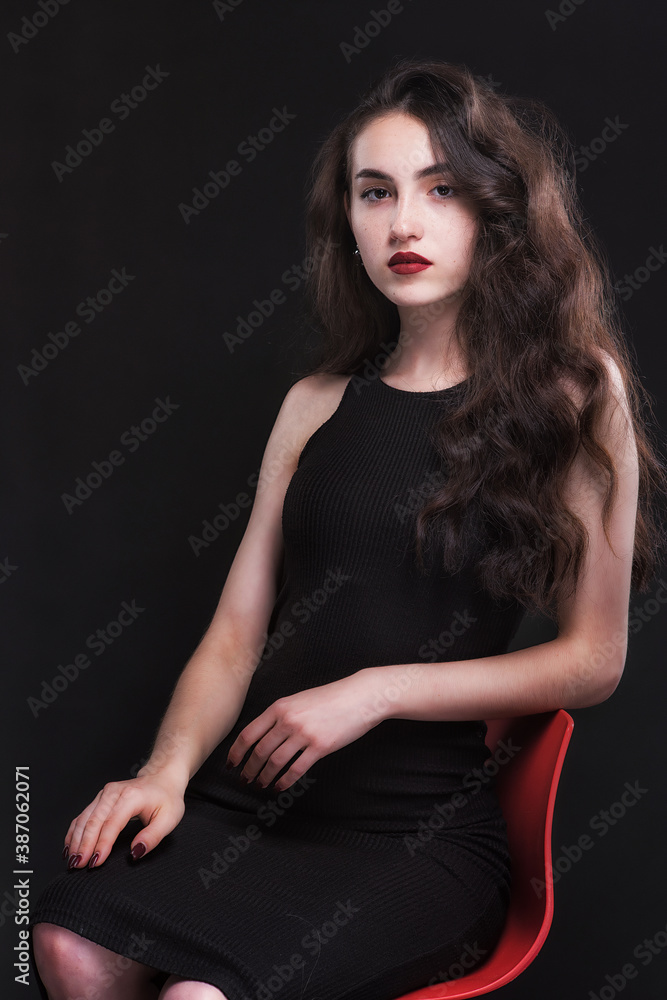 Beautiful slender young brunette with bright makeup in a black dress on a red chair