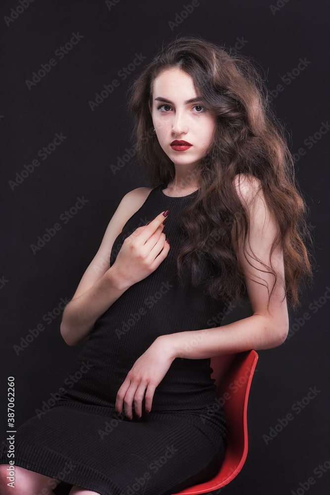 Beautiful slender young brunette with bright makeup in a black dress on a red chair