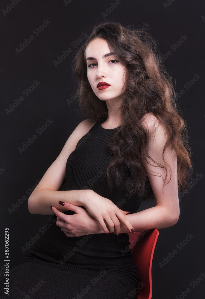 Naklejka premium Beautiful slender young brunette with bright makeup in a black dress on a red chair