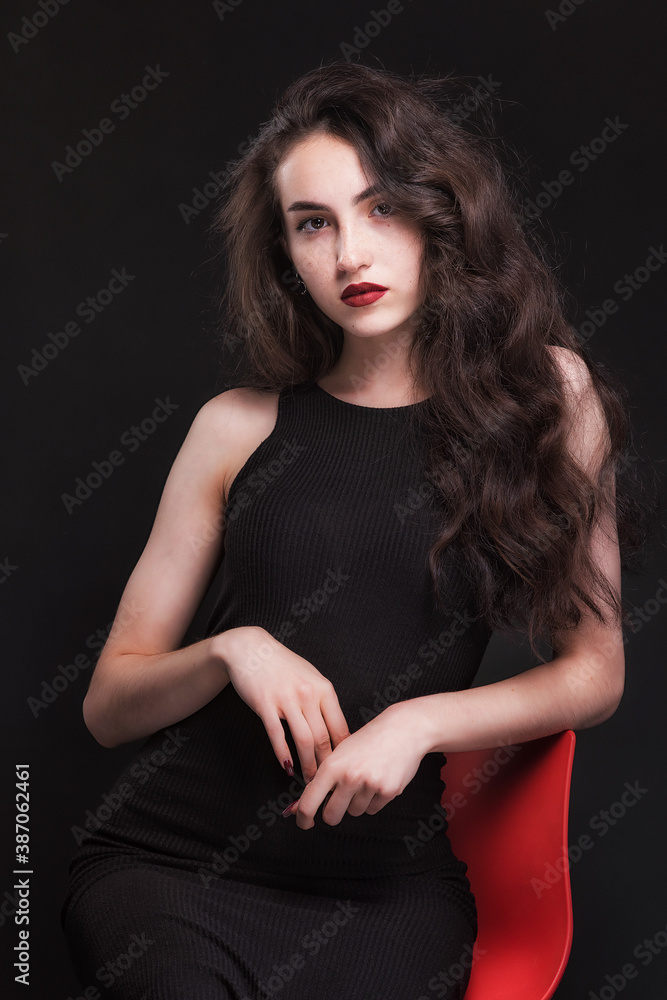Beautiful slender young brunette with bright makeup in a black dress on a red chair Beautiful slender young brunette with bright makeup in a black dress on a red chair