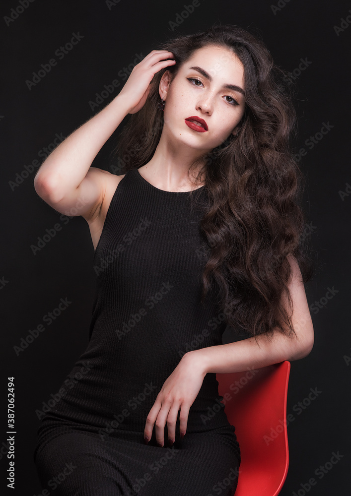 Beautiful slender young brunette with bright makeup in a black dress on a red chair Beautiful slender young brunette with bright makeup in a black dress on a red chair