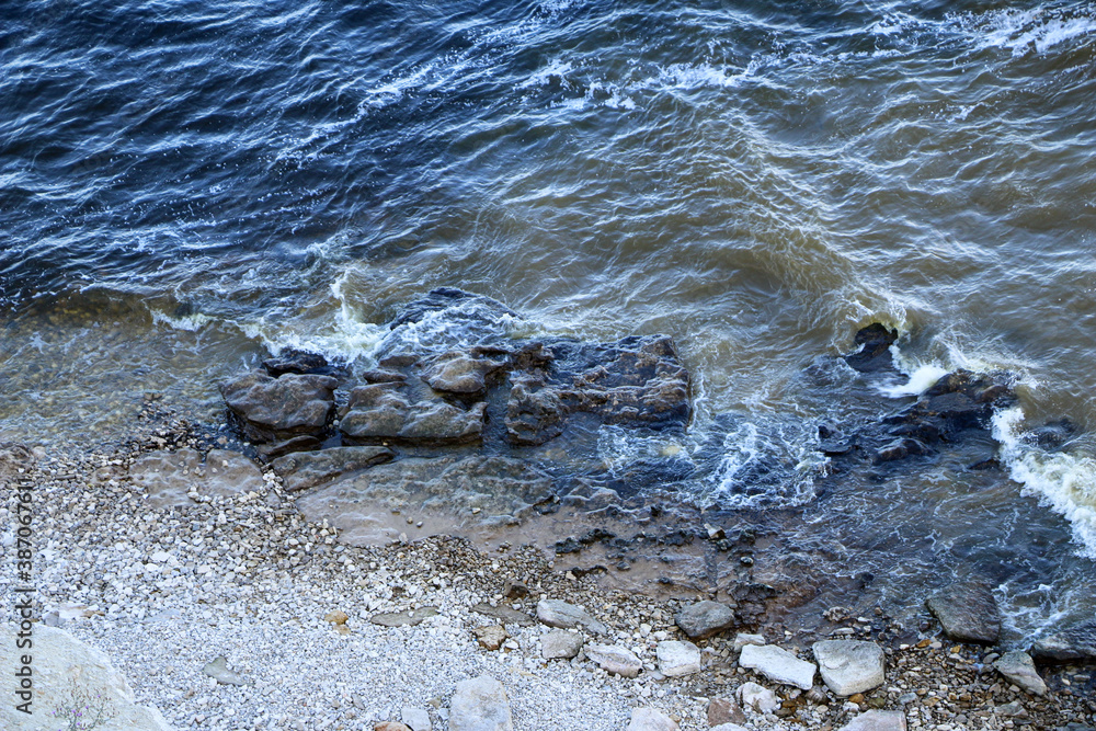 Obraz premium waves run onto the shore and crash against the rocks, creating many splashes and splashes near the shore. river surf in stormy weather near a stone pebble coast with foamy splashing waves.