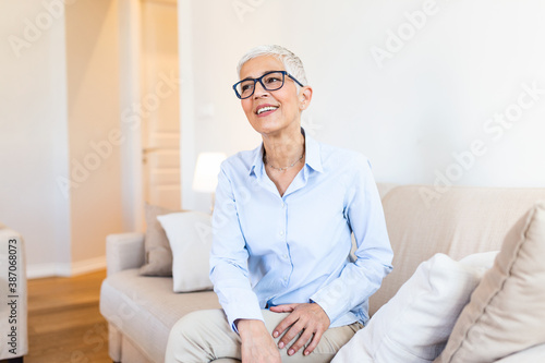 Older female with healthy smile sitting on couch at home. Smiling senior woman wearing glasses looking in distance.