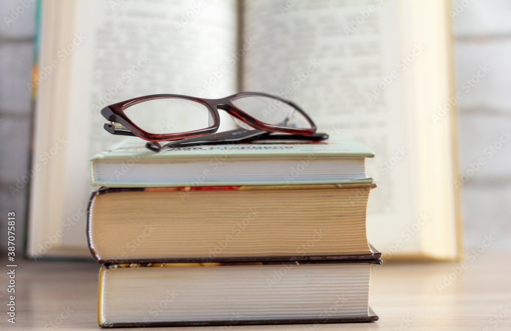 A stack of books with glasses and an open book in the background. A stack of books with glasses and an open book in the background.