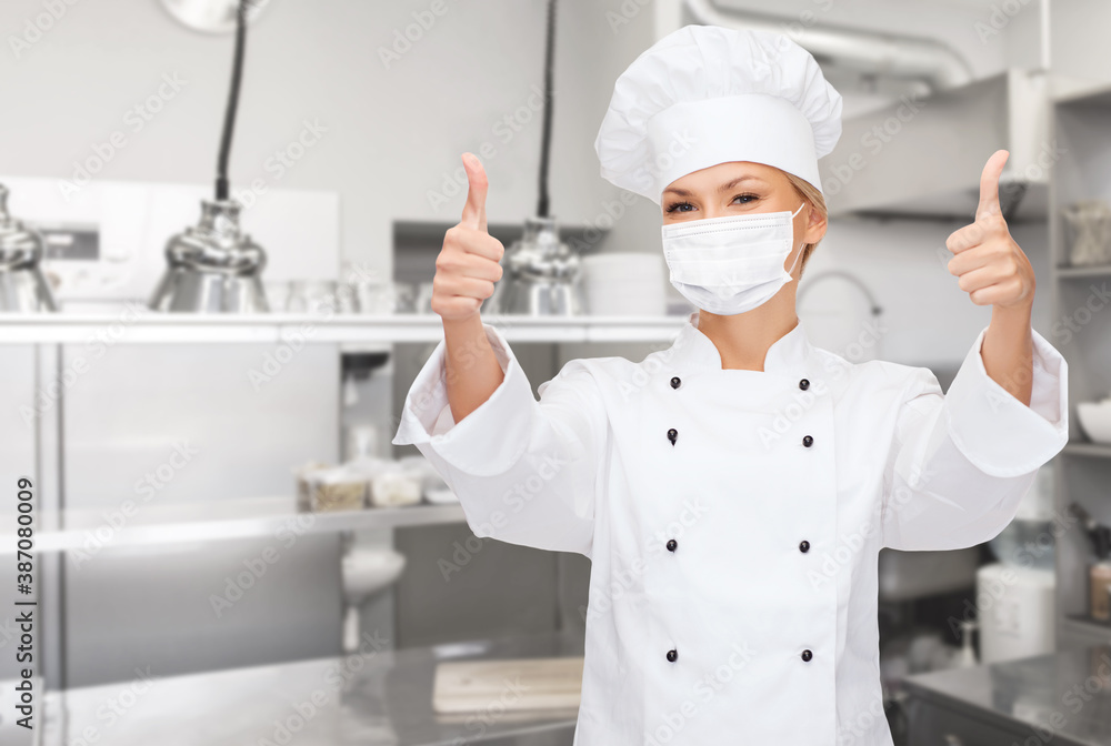 cooking, culinary and health concept - female chef in toque wearing face protective medical mask for protection from virus disease showing thumbs up over restaurant kitchen background cooking, culinary and health concept - female chef in toque wearing face protective medical mask for protection from virus disease showing thumbs up over restaurant kitchen background