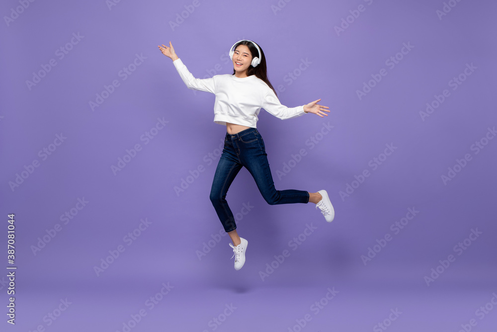 Young happy pretty Asian girl wearing headphones and jumping on isolated purple studio background Young happy pretty Asian girl wearing headphones and jumping on isolated purple studio background