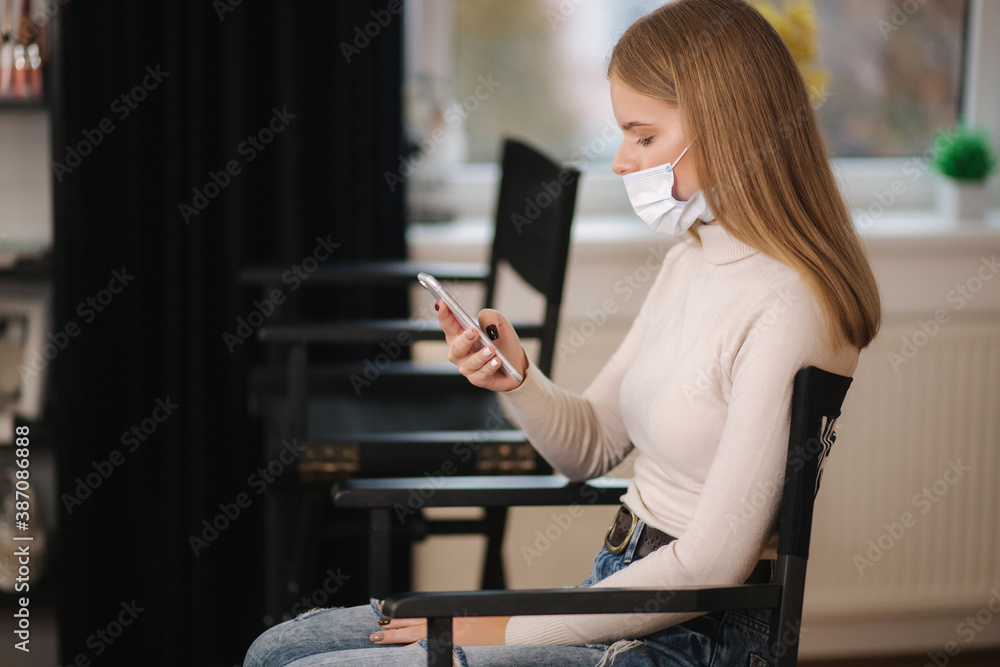 Female customer in beauty salon sitting on hight chair in medical mask and use phone. Beautiful young blond hair woman wiating for brow master. Open nose - obrazy, fototapety, plakaty 