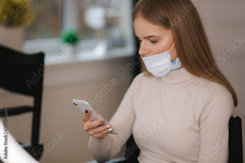 Female customer in beauty salon sitting on hight chair in medical mask and use phone. Beautiful young blond hair woman wiating for brow master. Open nose photo