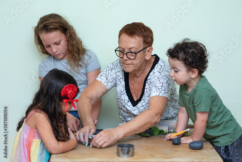 A Senior Woman Is Teaching A Little Kids How To Form With Clay. Grandmother Helping Little Grandchildren Sculpt Dough Figurines In The Home.