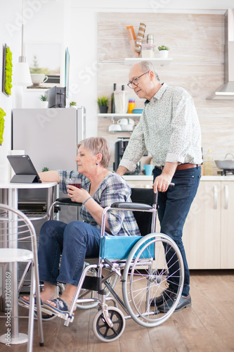 Handicapped senior woman in wheelchair browsing on tablet pc and talking with her husband in kitchen during breakfast. Positive mature man and woman.