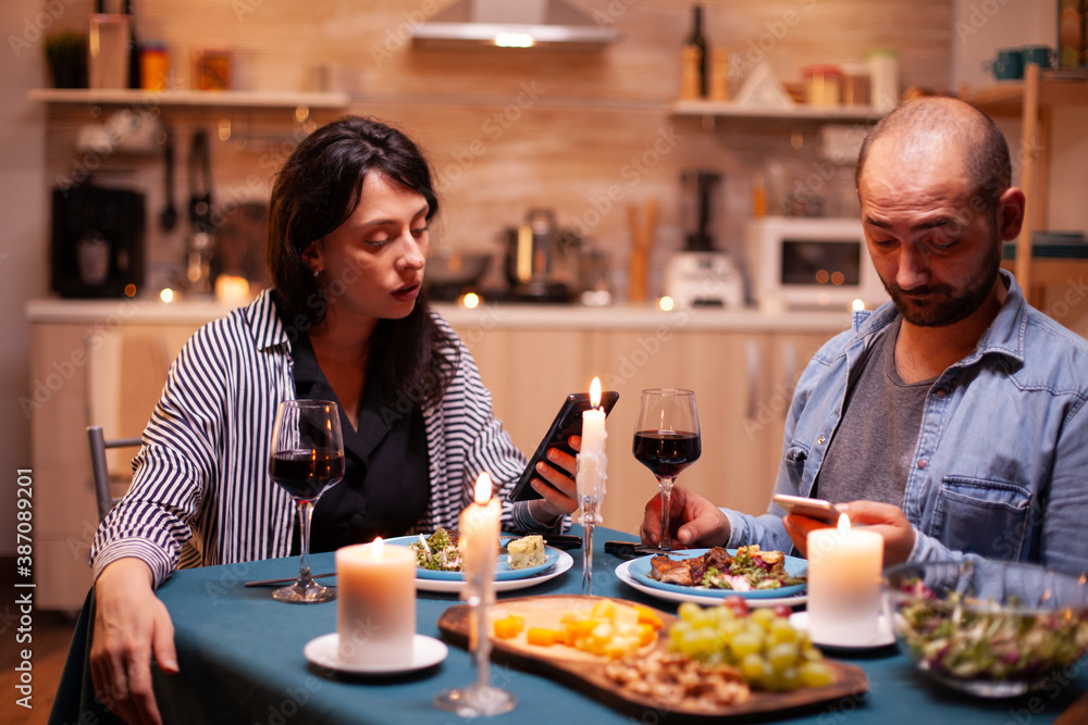 Happy couple watching video on smartphone in kitchen during anniversary. Adults sitting at the table in the kitchen browsing, searching, using smartphones, internet, celebrating anniversary.